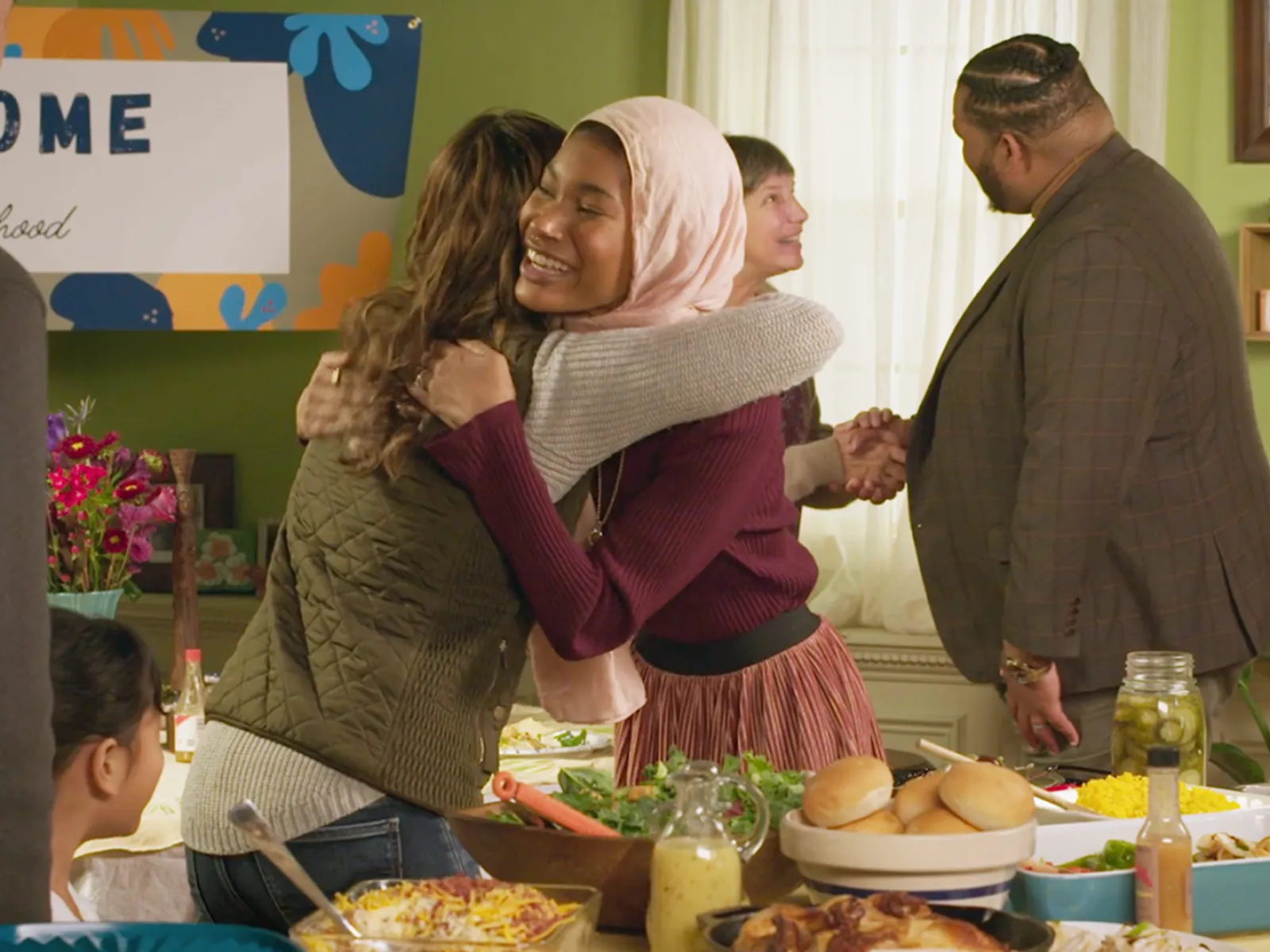 Two women giving each other a hug infront of a women and man shaking hands at a potluck.